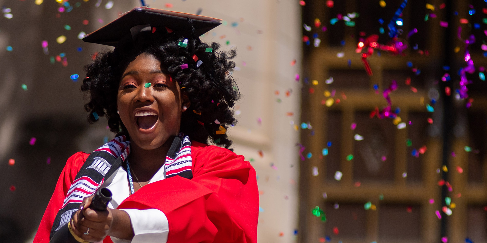 Ann-Lyssa Asare (Pardee’20) pops a champagne bottle full of confetti for a photo shoot in Marsh Plaza May 13, 2020. She wears a graduation cap and a red robe.