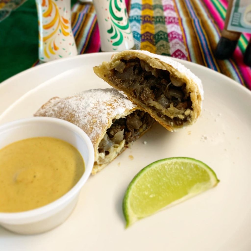 Photo of Carlos Olaechea’s Peruvian beef empanadas with rocoto chili sauce. Both rest on a plate with a lime wedge. Bright patterns are seen in the background.