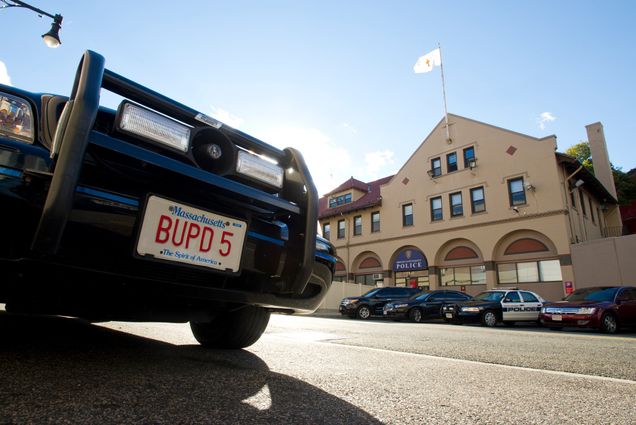 Image of the license plate of a BUPD vehicle. The plate reads 'BUPD 5.' A station flying a flag and other police vehicles are seen in the distance.