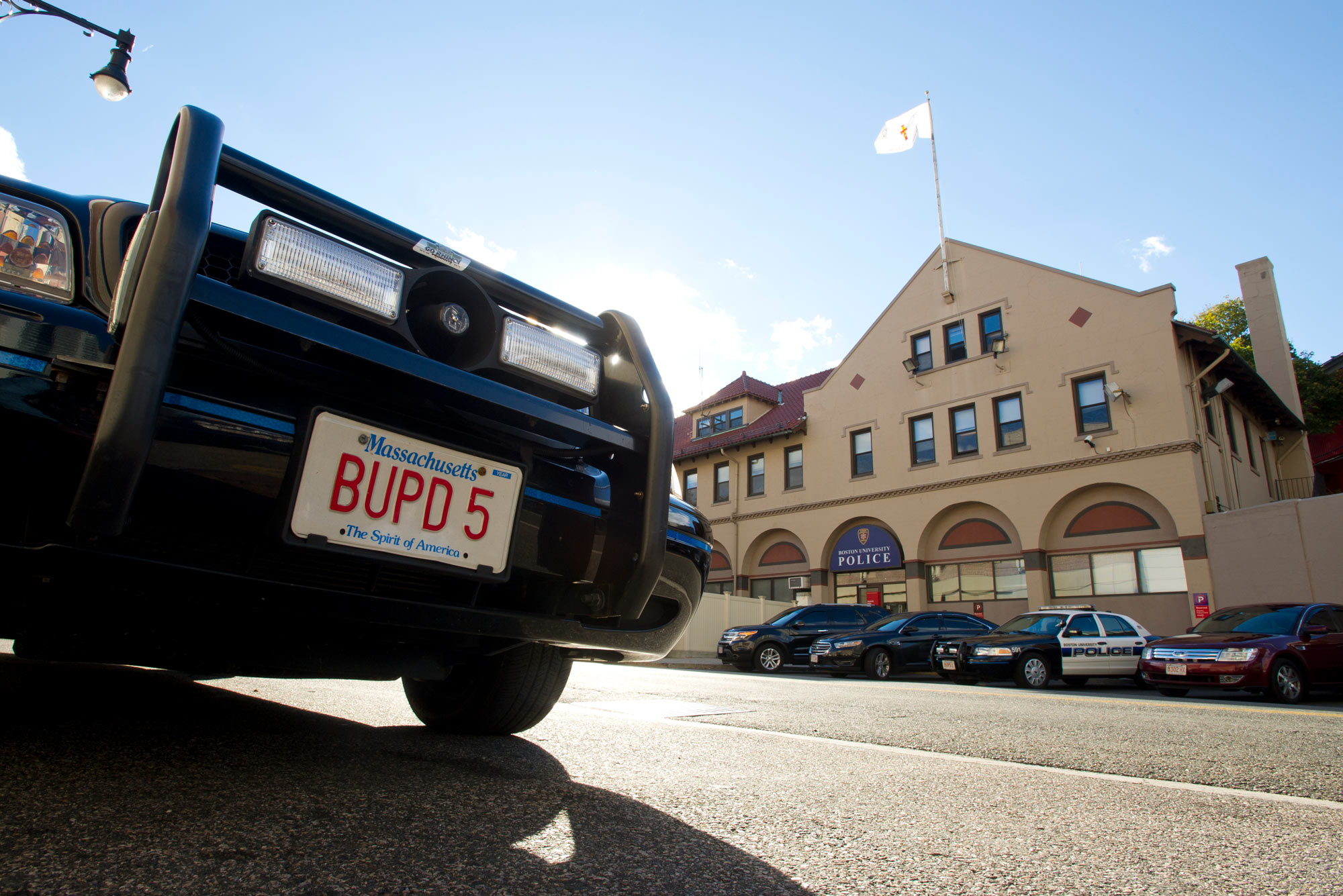 Image of the license plate of a BUPD vehicle. The plate reads 'BUPD 5.' A station flying a flag and other police vehicles are seen in the distance.