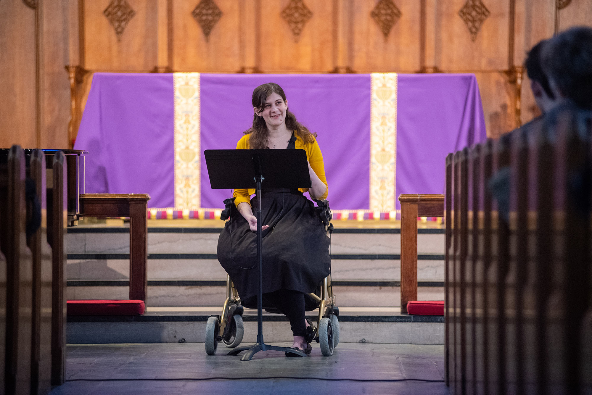 Elizabeth Foster (STH’22), who has a condition that makes prolonged standing difficult, preaching from her walker chair during a Marsh Chapel service dedicated to those with disabilities. An altar with a purple cloth is seen behind her.