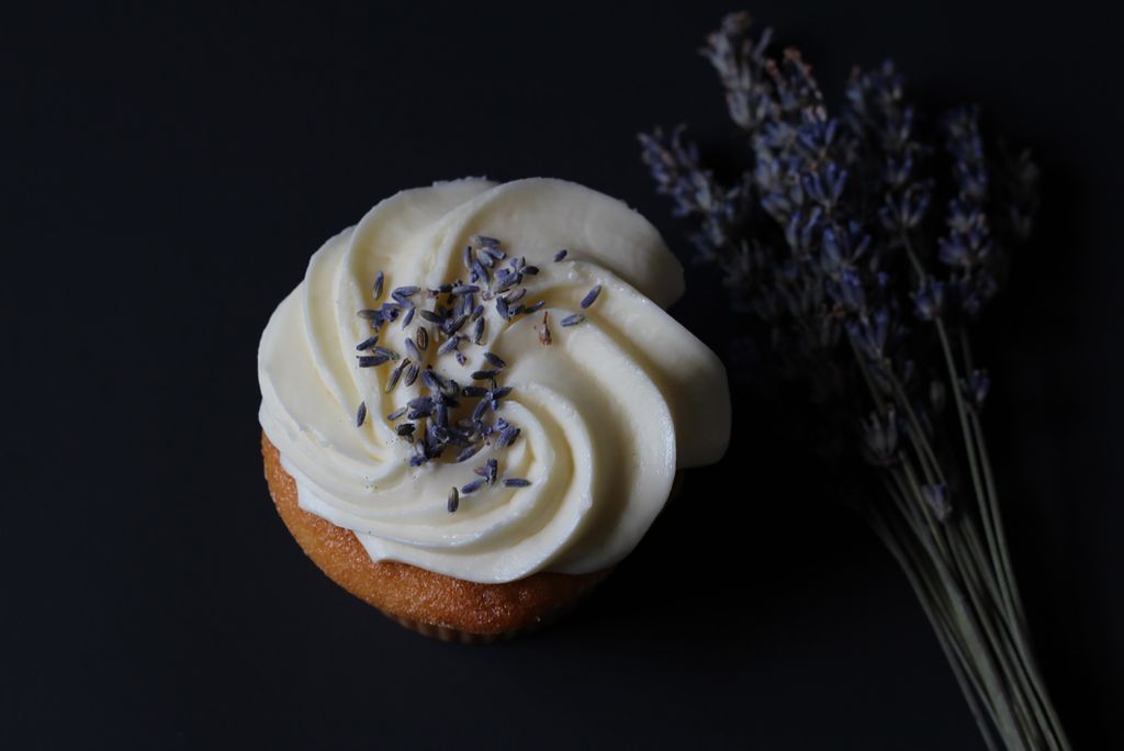 Photo of a lavender cupcake. The cupcake is vanilla with white frosting, and lavender flowers are sprinkled on top. The image is top lit and the background is dark.