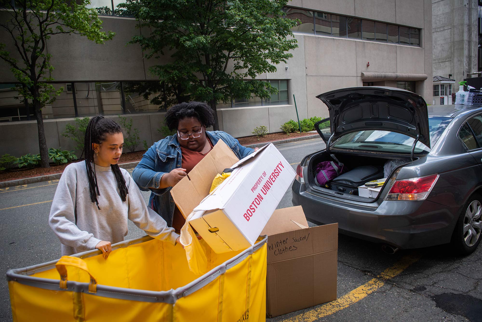 Photo of Katerina Chew (left) (MET 20) and Robyn Volcy-Lee (SAR 20, SPH 21) pack up their car on June 1, 2020. Students were able to return to campus to pick up their belongings. Volcy-Lee holds a "Property of Boston University Box" as Chew holds a yellow moving cart.