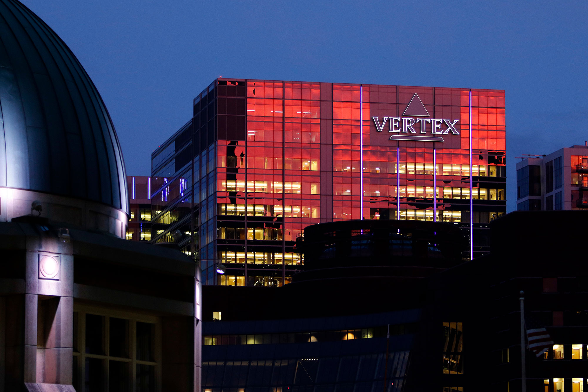 The sunset reflects off the Vertex Pharmaceuticals headquarters building, Thursday, Sept. 21, 2017 in Boston's Seaport. A sign on the building reads 'Vertex' in purple. The top dome of a building is seen in the bottom left foreground.