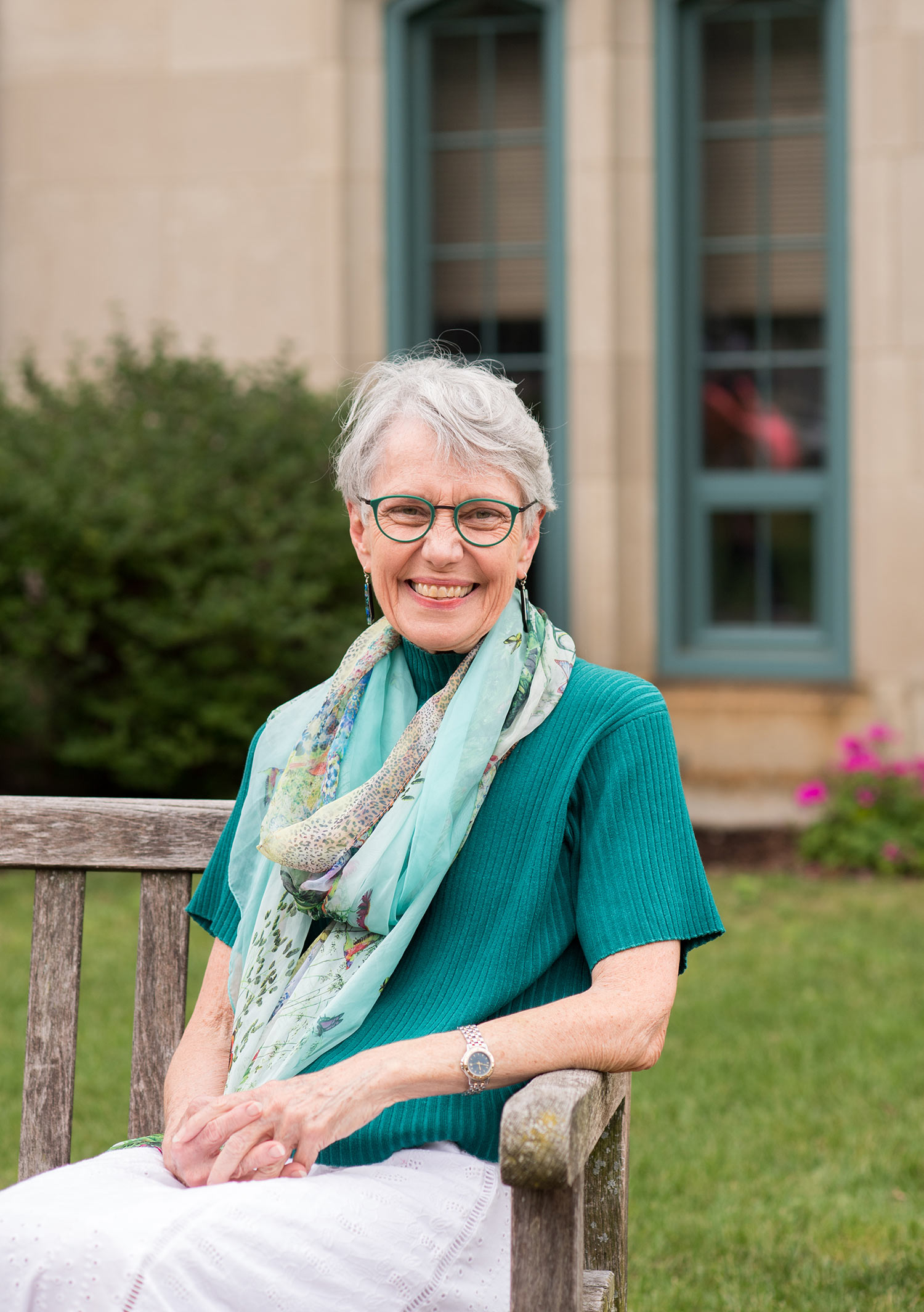 Photo of Dean Moore sitting on a bench in a scarf and teal shirt. Flowers and a grass patch are seen behind her.