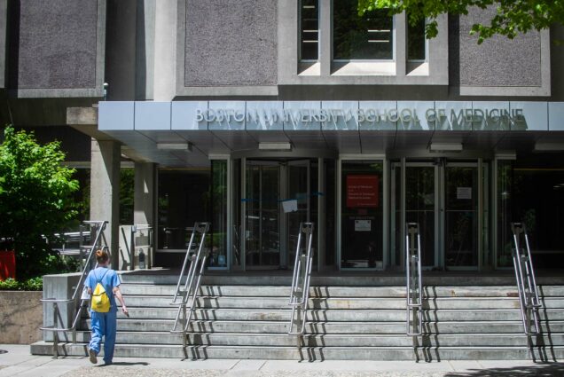 Photo of the exterior of the Boston University School of Medicine on Tuesday, May 19, 2020. A person in scrubs wearing a small yellow backpack walks up the stairs towards the front door.