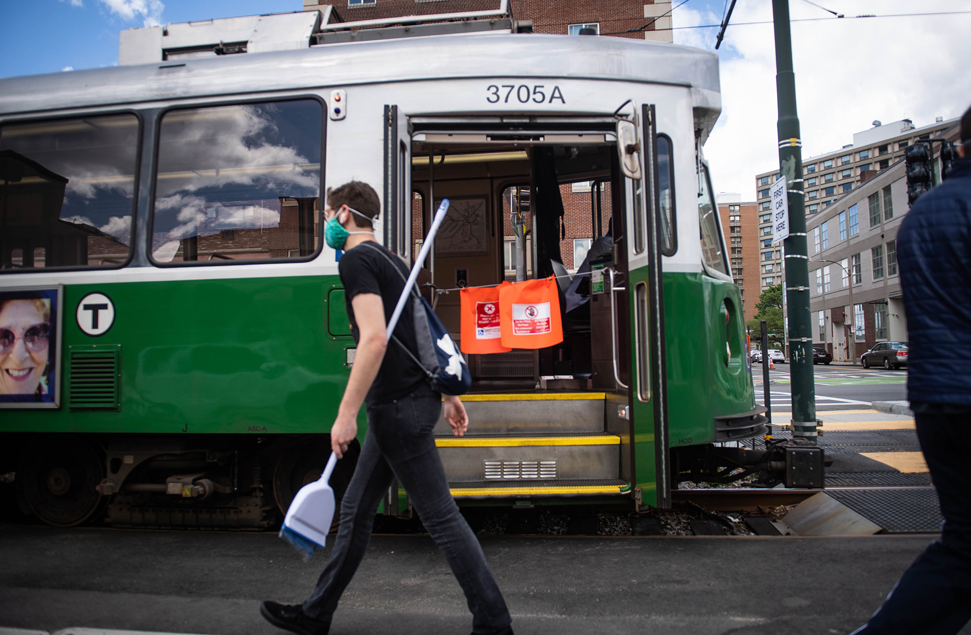 A student in a mask holding a broom walking by a green line train on Comm Ave