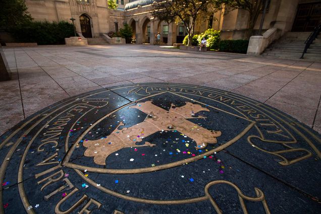Photo of the BU Seal with colorful confetti on the ground. Two people walking on Marsh Plaza are seen in the distance.
