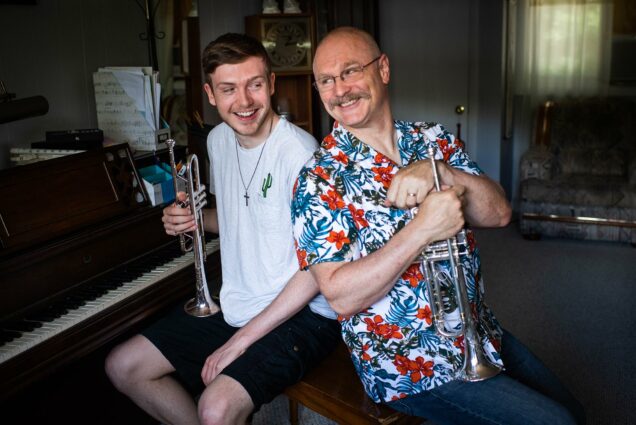 Photo of Terry Everson, the primary instructor of trumpet performance in CFA's School of Music and how son, Peter pose for a photo at their Framingham home on June 19 ,2020. Terry wears a bright blue and red floral shirt and holds a trumpet, leans and looks towards his son who wears a white shirt and holds a trumpet as well.