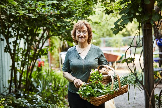 BU alum Renee Shepherd, founder of seed company Renee’s Garden, at her test gardens in Felton, California. Renee wears a green shirt and holds a basket of herbs and what looks like onions or garlic. A wheelbarrow and other garden equipment are seen blurred behind her.