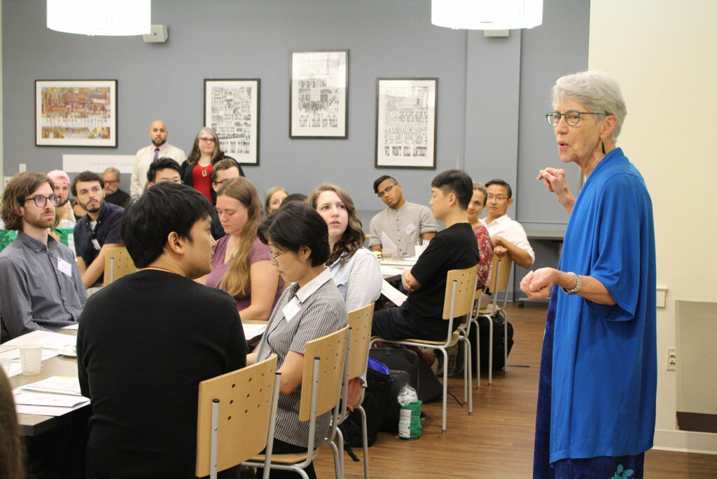 Dean Moore in a light blue sweater is seen addressing students during 2018 orientation. Students turn in their seats to listen to her speak.