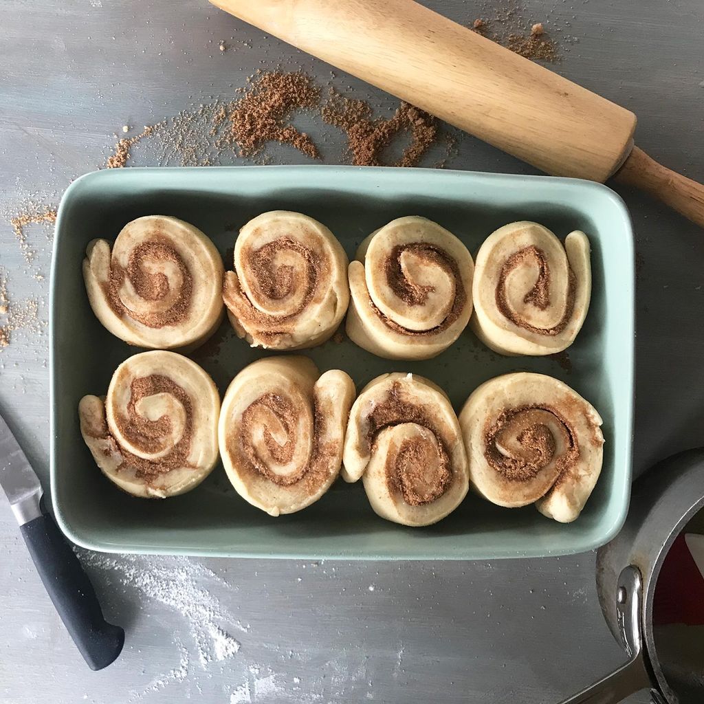 Aerial photo of eight cinnamon rolls before being glazed and baked. Around the tin a knife, rolling pin and sugar are scattered.