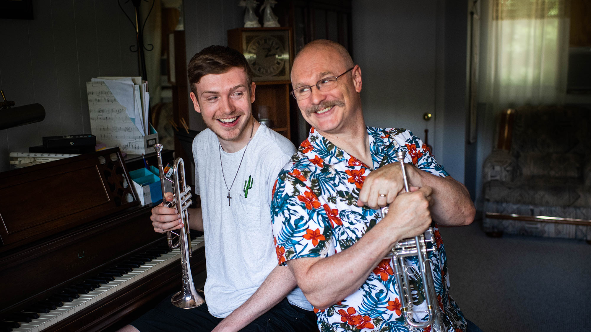 Photo of Terry Everson, the primary instructor of trumpet performance in CFA's School of Music and how son, Peter pose for a photo at their Framingham home on June 19 ,2020. Terry wears a bright blue and red floral shirt and holds a trumpet, leans and looks towards his son who wears a white shirt and holds a trumpet as well.