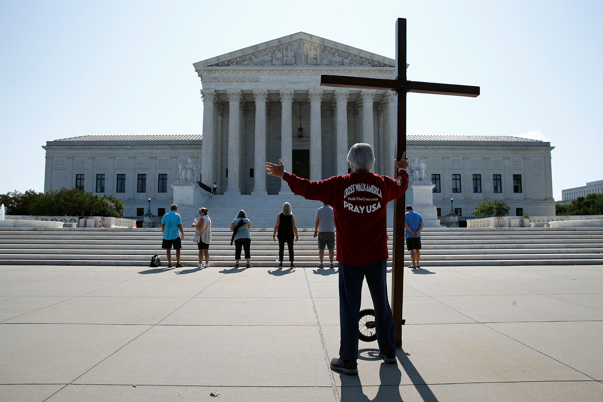 Photo of a man holding a cross and in the background, a group of people praying outside the Supreme Court on July 8, the day the court handed down decisions giving religious groups broad discretion in firing employees and denying contraceptive coverage. Behind the Supreme Court building the sky is clear and sunny.