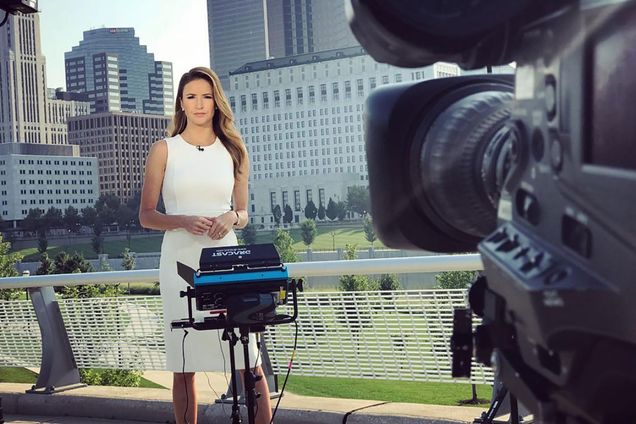 Photo of Kristin Fisher behind a light and a camera in a white dress reporting with a city skyline behind her.