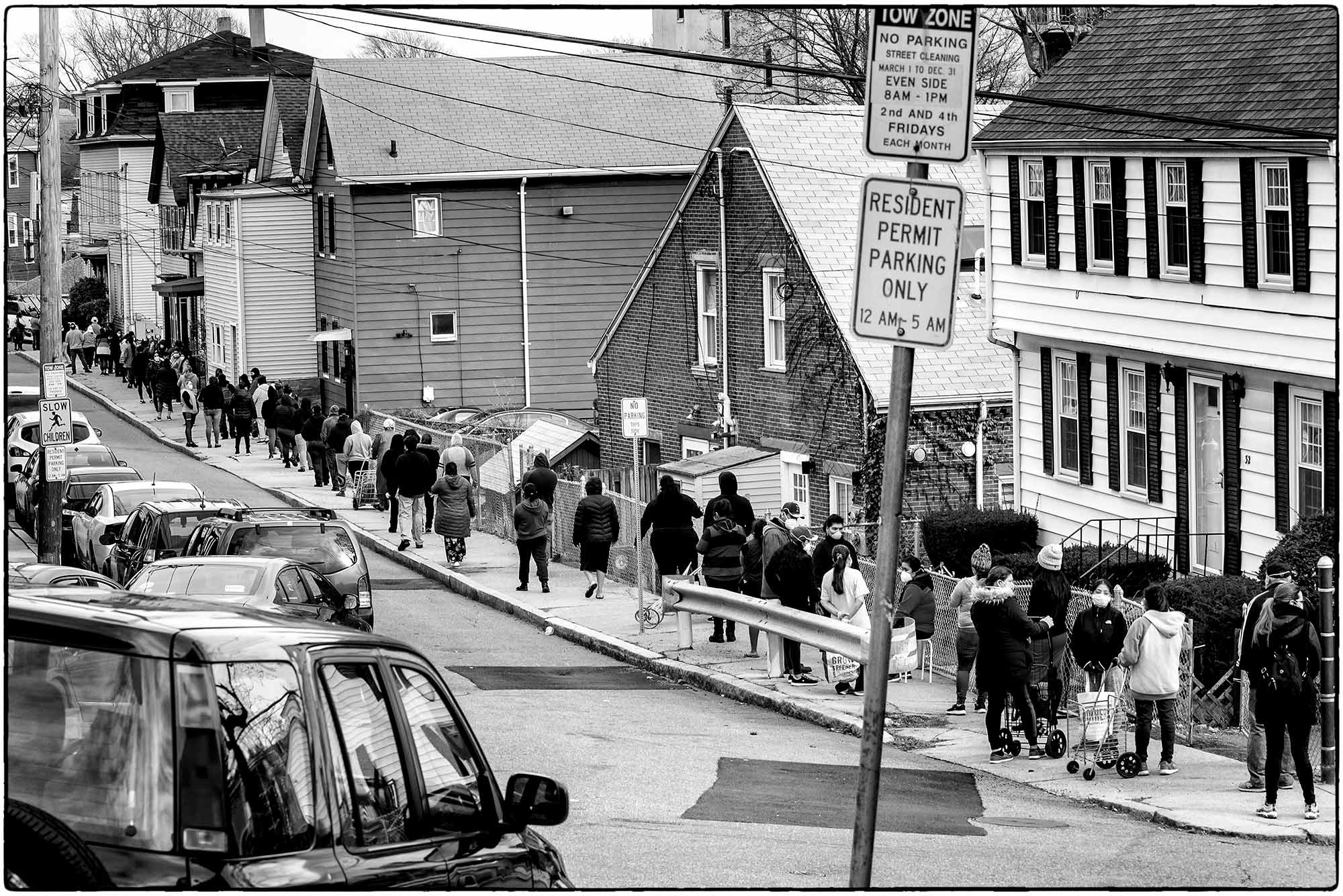 Photo of a group of Chelsea, Mass. residents lined up, many in sweatshirts or jackets, to pick up food at a food pantry. The photo shows a long line on the other side of the street, with a car and a "Resident Parking Only" Sign in the foreground. The photo is black and white.