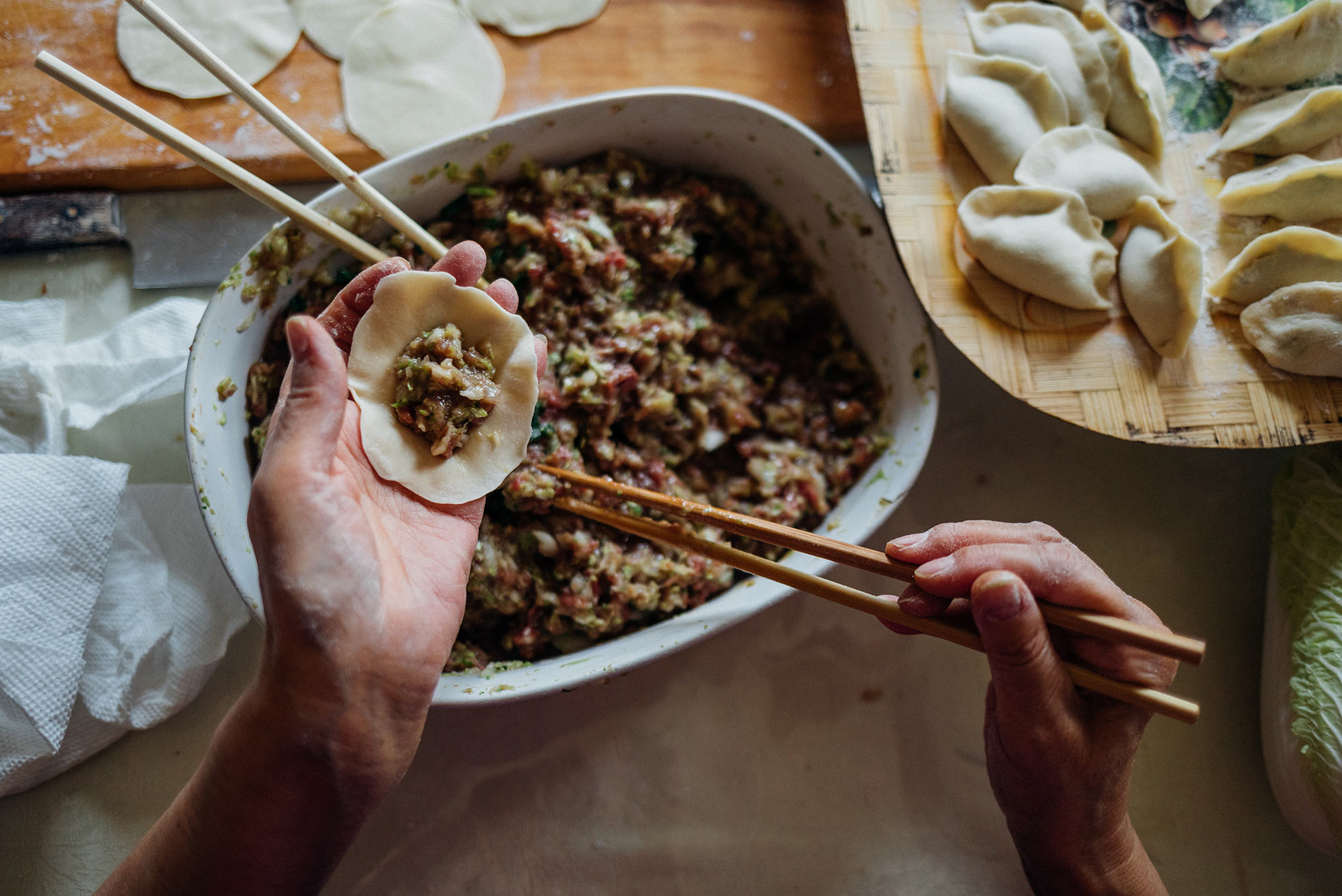 Photo of someone making dumplings. In one hand, they hold a dumpling wrapper, in the other, a pair of chopsticks that they use to add filling to the wrapper. A bowl of filling, empty wrappers, and finished dumplings are seen in the background.