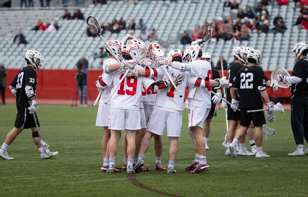 Men’s lacrosse players, dressed in white jerseys, celebrate in a huddle during a game.