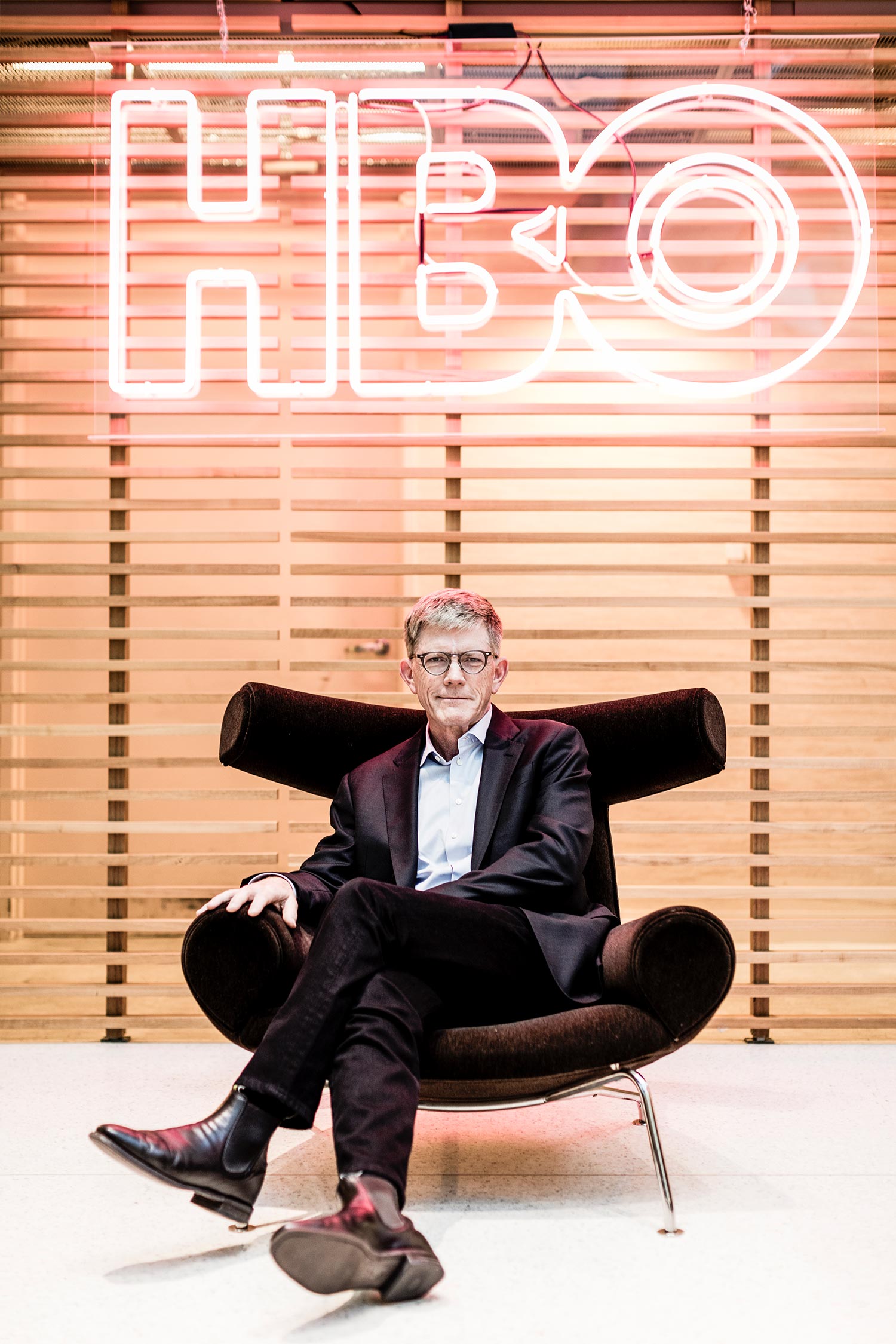 Portrait of Jay Roewe (COM’79) in a black suit in a stylish black chair. Behind him, a neon sign reads "HBO"