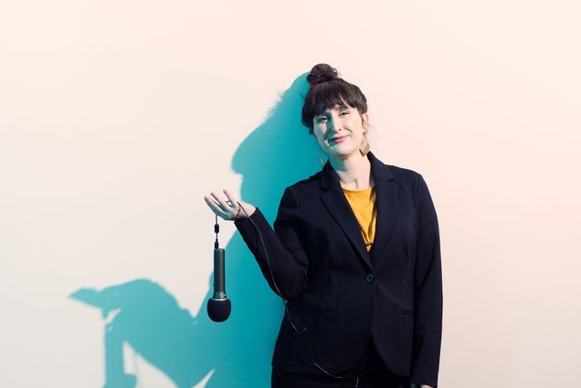 Portrait of Erika B. Hess (’09), host of the podcast I Like Your Work, standing with a microphone between her fingers against a wall. A deep blue shadow silhouette is seen behind her.