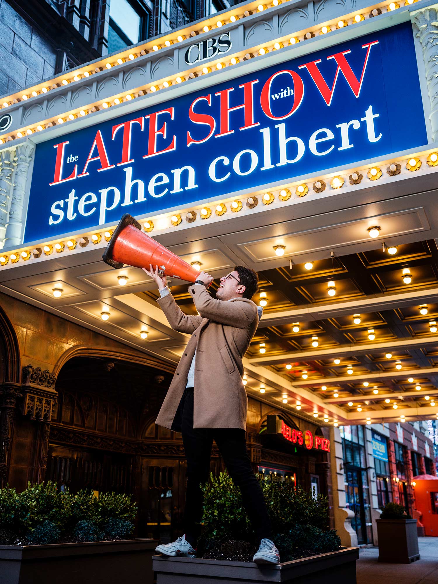 Felipe Torres Medina, a staff writer for The Late Show with Stephen Colbert, poses outside of the Late Show sign and yells into an orange traffic cone as if it were a megaphone.