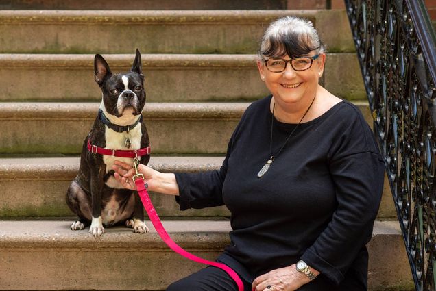 Maureen Mahoney, pictured here with Boston Terrier Augie, is retiring after 26 years at BU as a crisis counselor, helping people traumatized by sex asssault, domestic violence, hazing, fires, accidents, and deaths. Mahoney and Augie sit on stairs.