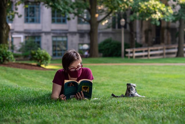 Sydney Kovar, who works in the Learning and Event Technology Services department relaxes during her break on BU beach in the early evening July 9, 2020. She lays on the grass reading a book by Toni Morrison, her shoes next to her on the left. A tree and grassy space are seen behind her.