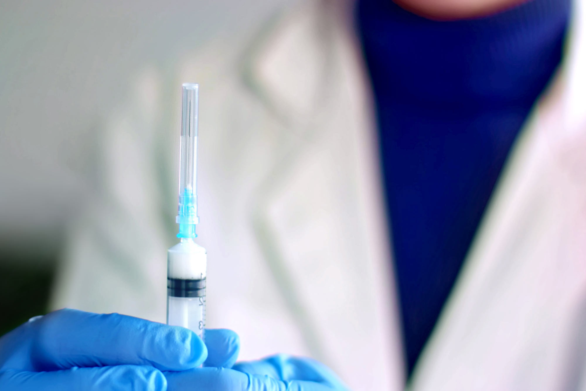 A photo of a doctor in a white lab coat and blue gloves holding up a capped syringe