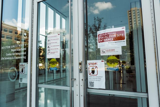 Photo of the glass door at the entrance of the Rajen Kilachand Center at Boston Univeristy. Paper signs on the door read “COVID-19 Testing Collection Site Entrance” and “Stop! Have you completed your required daily symptom survey?”