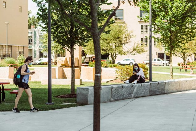 Photo of two students outside of Covid-19 testing lab at Kilachand Center with masks on. One sits on a bench and looks at their phone, the other walks by with a backpack.