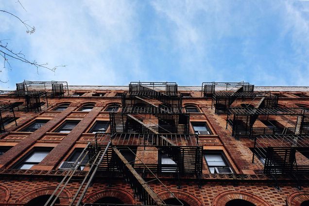 Photo of an apartment building in Williamsburg, NY with a brick facade and a series of black fire escapes from the ground level looking up. A blue sky with a few clouds is seen behind the building.