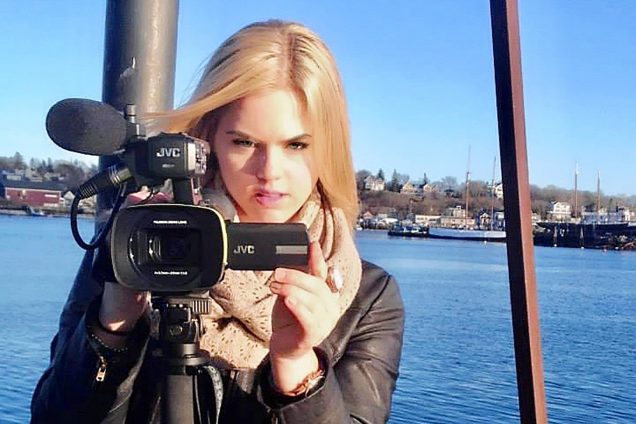 Photo of Victoria Price holding a video camera and recording during her time at BU. Behind her a body of water and shoreline are seen. Price wears a black leather jacket and scarf.