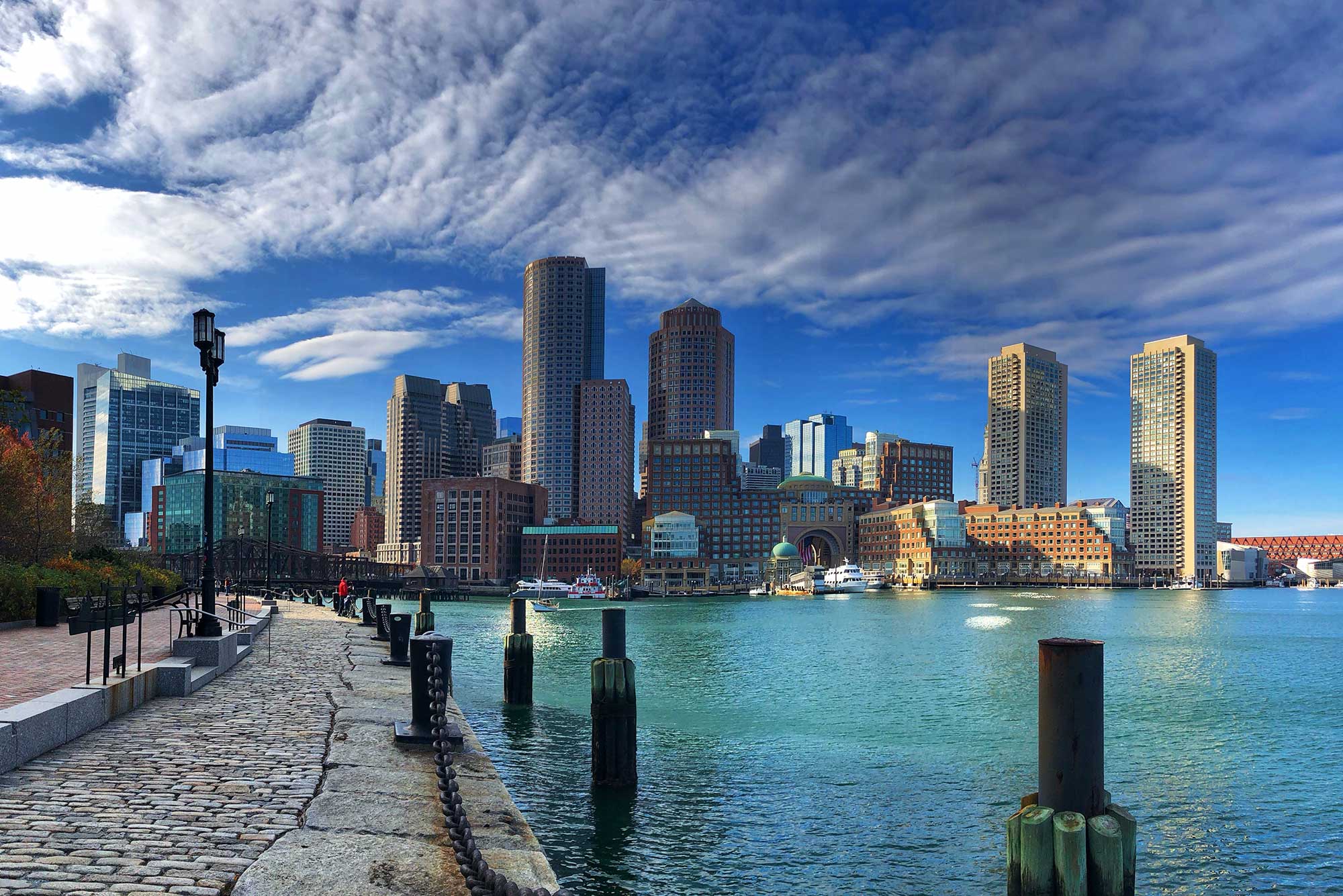 Photo taken from Boston Seaport looking towards the Boston skyline. The water is light blue, and the sky is blue with some clouds.