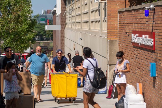 Photo of students and their families pushing yellow carts and stopped on the sidewalk with their belongings along Babock Street during move in 2021. Some folks are seen with masks on. A BU sign on a building is seen in the background.