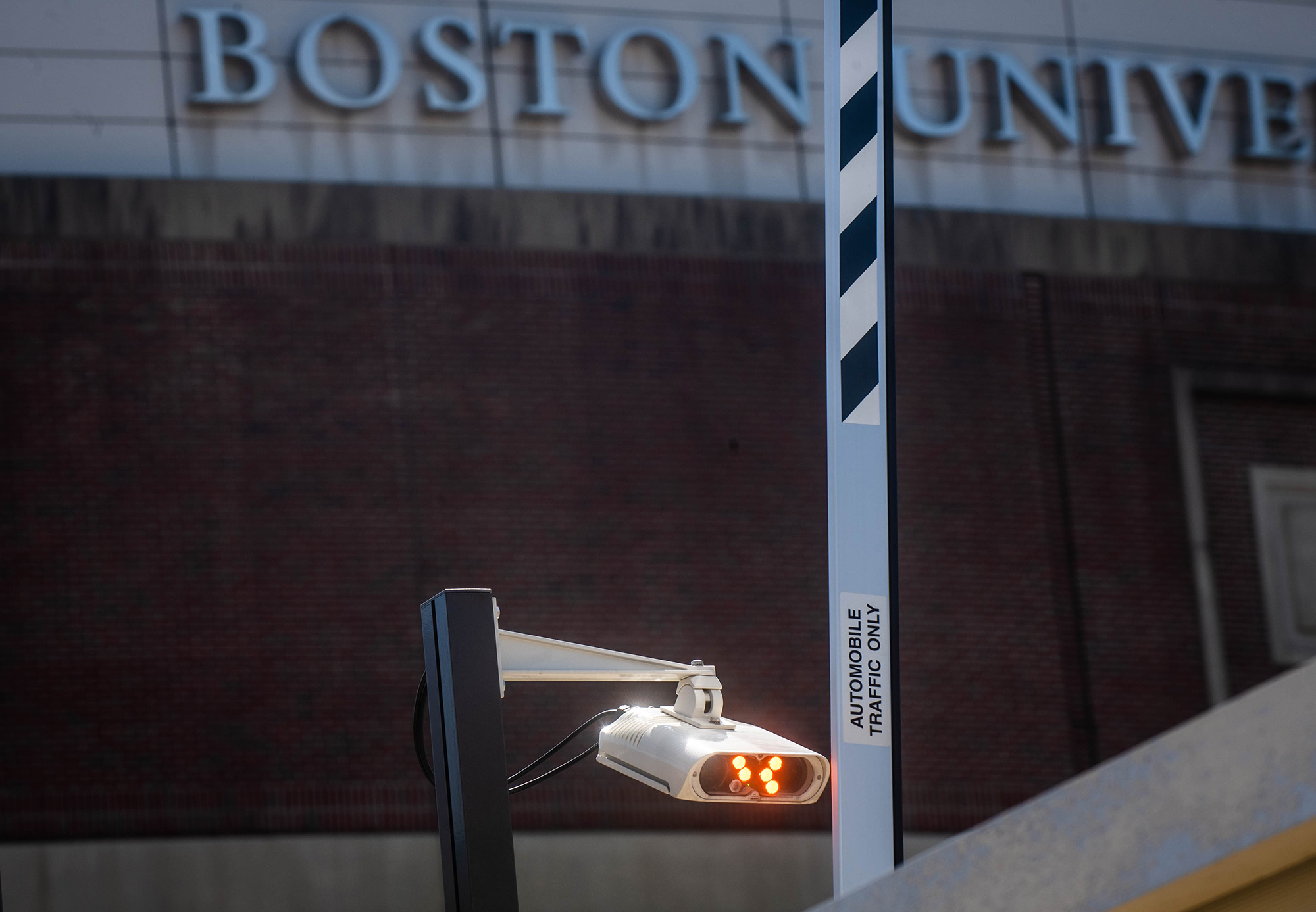 New cameras are seen at the Agganis Arena parking garage on August 3, 2020. The new parking program will read license plates instead of stickers or hang tags. A camera with orange lights hands next to a parking lot arm. A Boston University sign is seen in the background.