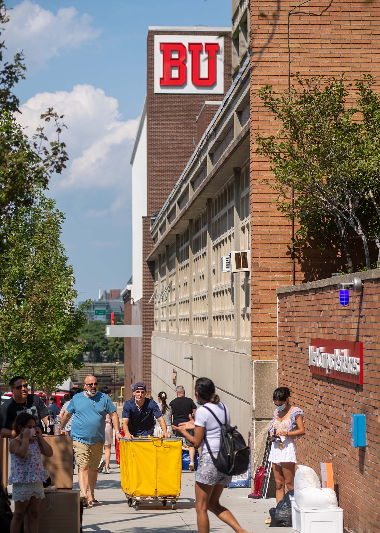 Photo of students and their families pushing yellow carts and stopped on the sidewalk with their belongings along Babock Street during move in 2021. Some folks are seen with masks on. A BU sign on a building is seen in the background.