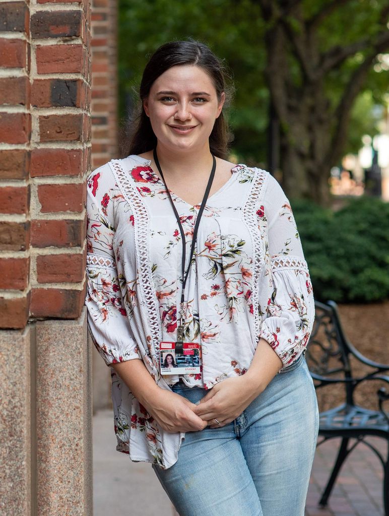 Jori Chambers wears a lanyard with her BU ID, a flowery blouse and jeans, and leans against a brick wall. A bench and trees are seen in the background.