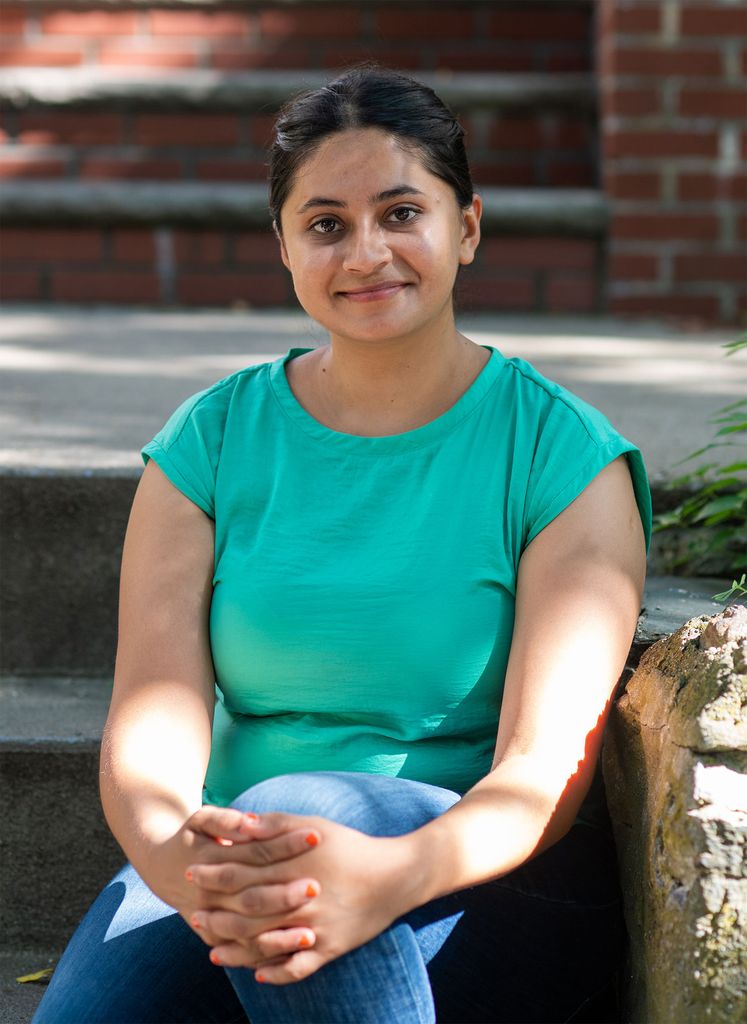 Fatima Aqeel sits on steps near her Allston home on July 28. Aqeel rests her hands on her knees, wears a teal blouse and jeans, and smiles.