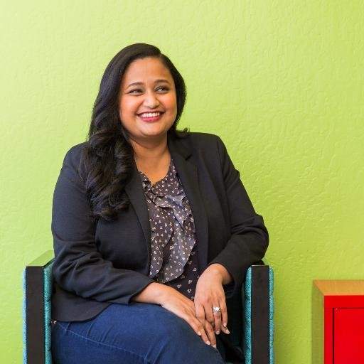 Photo of Kamakshi Sivaramakrishnan (ENG ’00)  in a blazer and jeans sitting in a chair in front of a green background. 