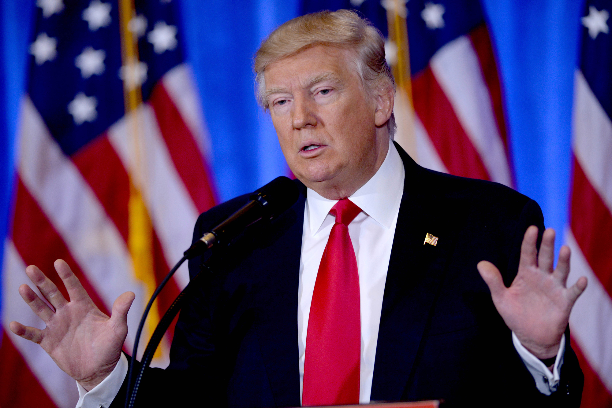 A photo of Donald Trump speaking at a podium with his arms raised. A row of American flags are behind him.