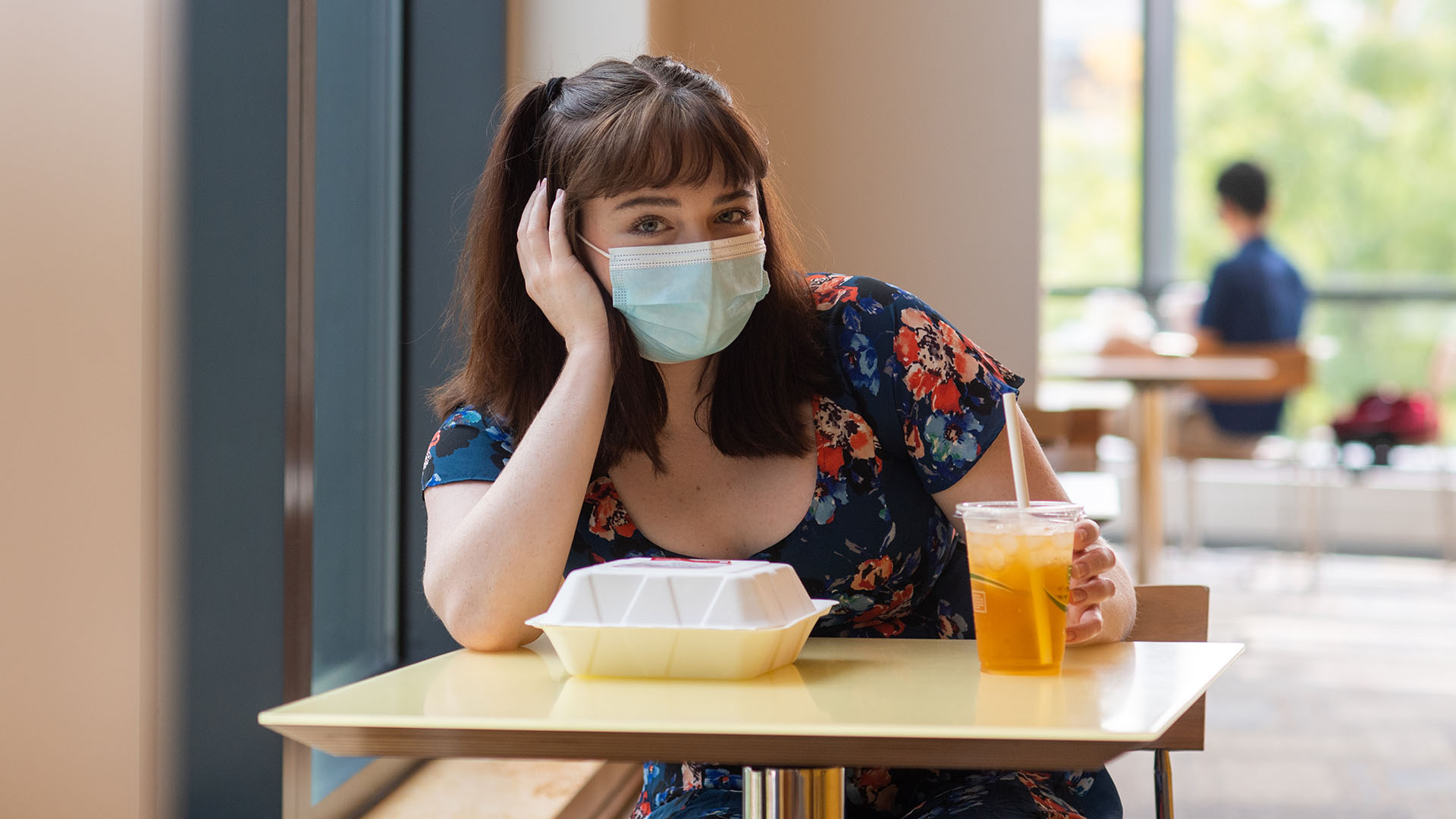 Photo of Brigid Kane (COM’22) sitting at a small dining hall table by herself with a face mask on. A paper to-go container and drink sit in front of her; she rests her hand on her face. Overlay has a video play button.