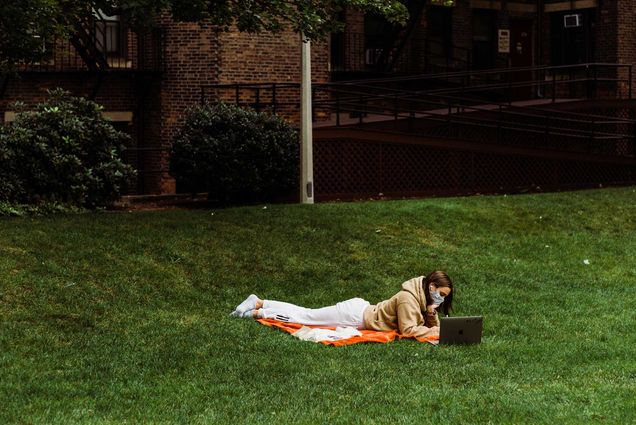 Photo of a student in a sweatshirt laying down in the grass at BU Beach in a mask. The student works on their laptop.