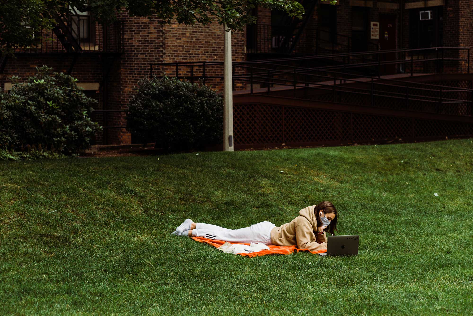 Photo of a student in a sweatshirt laying down in the grass at BU Beach in a mask. The student works on their laptop.