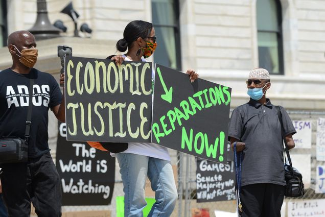 Photo of Juneteenth reparations rally to demand reparations from the United States government. A rally-goer holds two signs, one says "economic justice" while the other says "reparations now!" Other masked individuals are seen in the frame.