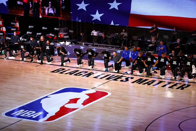 Photo of Players from Orlando Magic and Milwaukee Bucks who took a knee during the national anthem before the start of Game 3 of an NBA basketball first-round playoff series, Saturday, Aug. 22, 2020, in Lake Buena Vista, Fla. The players where black lives matter t-shirts. The court parquet floor reads "Black Lives Matter" and has a large NBA logo. The American flag is seen behind the kneeling players.