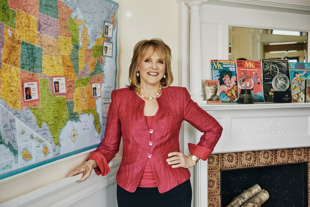 Photo of Barbara Lee in a bright red pant suit posing in her study near a map of woman she has helped elect to prominent roles in the government. Behind her on a mantle sits a few issues of the magazine Ms.