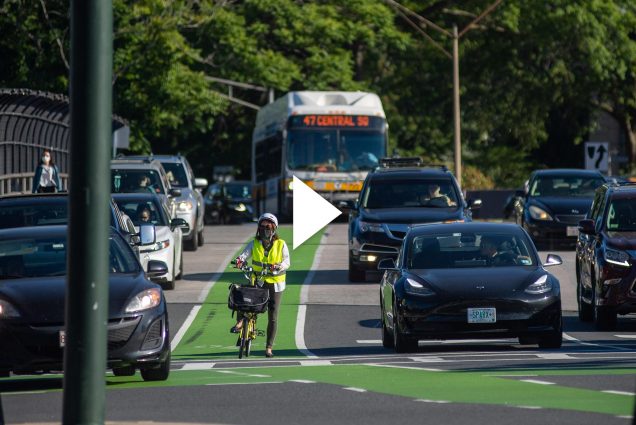 Photo of a person in a green vest and helmet on a bike waiting in a bike lane at a light. Many cars and city buses are seen around them. Overlay has video play button.