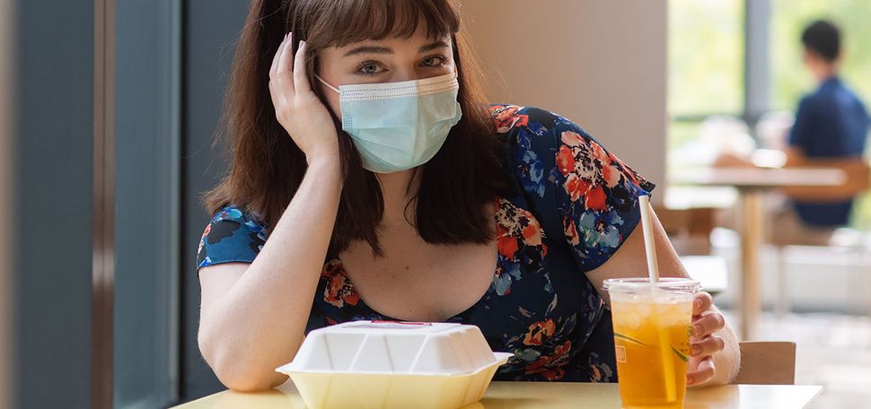 Photo of Brigid Kane (COM’22) sitting at a small dining hall table by herself with a face mask on. A paper to-go container and drink sit in front of her; she rests her hand on her face. Overlay has a video play button.