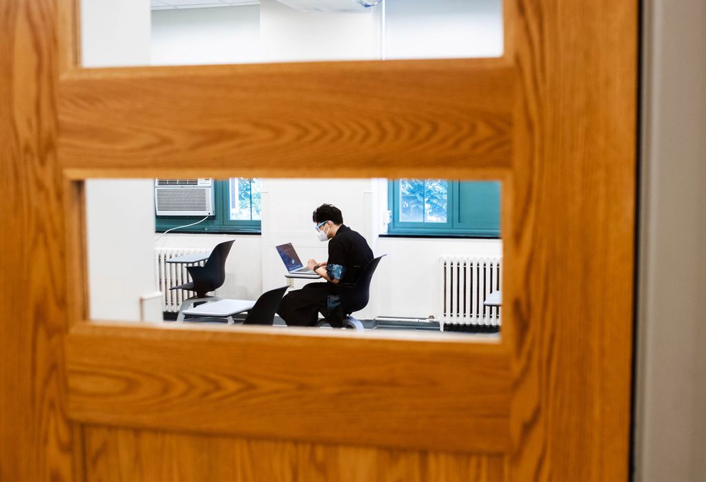 Photo from outside a classroom looking in through the door's window; inside, a lone student sits with a mask and looks at their laptop. This photo was taken at the College of Arts & Sciences on the first day of classes.