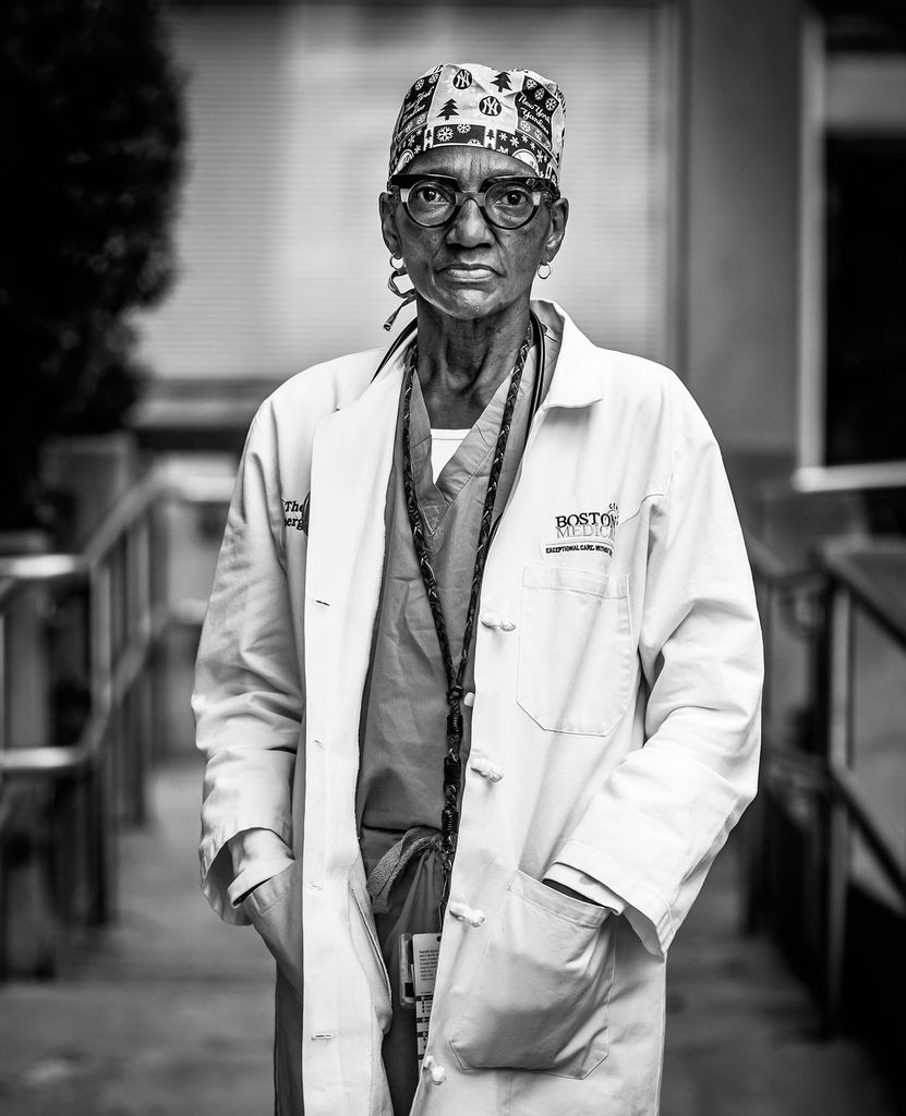 Black and white portrait of  Thea James, BMC associate chief medical officer and vice president of mission, and MED associate professor of emergency medicine. Dr Thea wears a white lab coat and stands with hands in their pockets. 